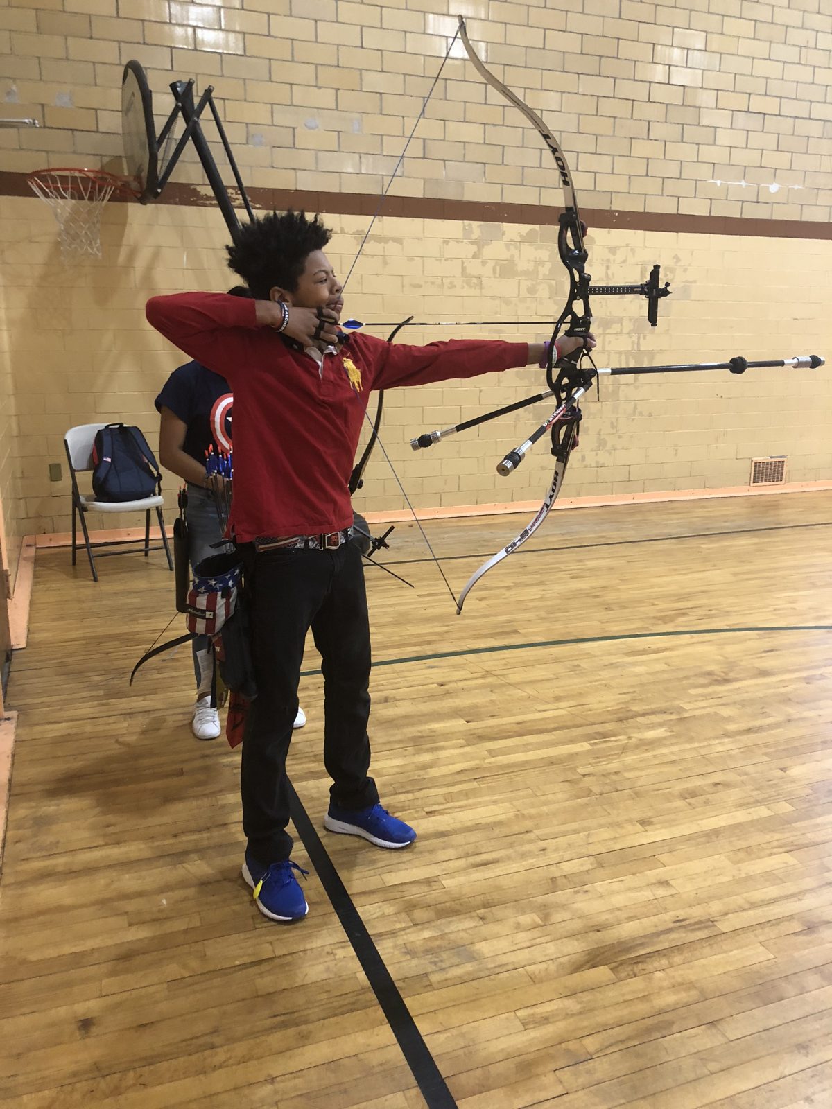 A young archer in a red polo at full draw with a recurve bow in a school gym